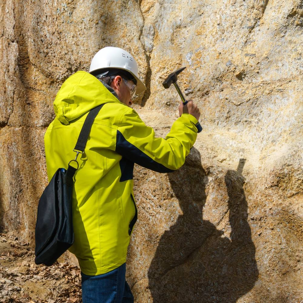 Geologist in a helmet beats a rock with a hammer to take a sample of the mineral